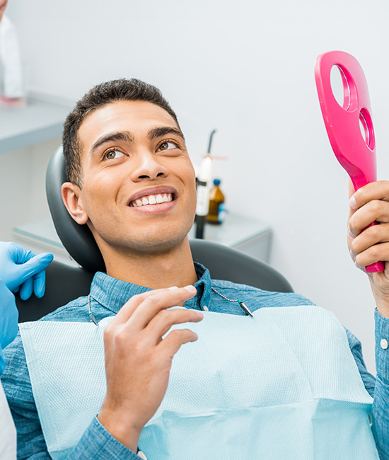 A dentist is smiling at the camera while sitting in a dental chair, holding a pink toothbrush.