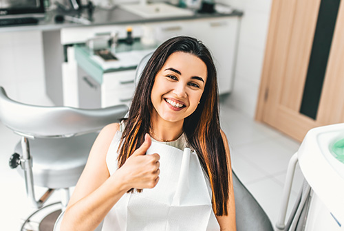 The image shows a smiling woman in a dental office, holding up a thumbs-up gesture with her right hand.